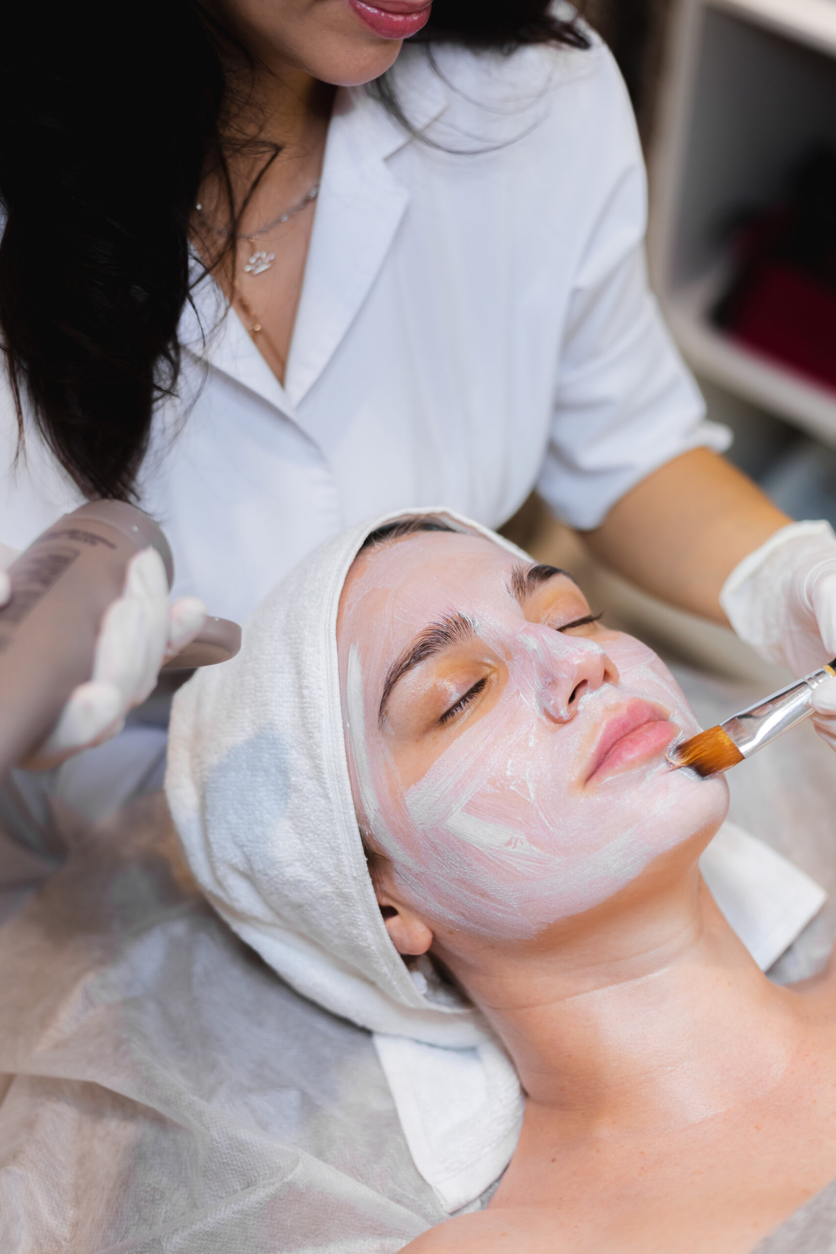 Skin behanldungh applies a white moisturizing mask to the face of a young girl client in a spa beauty salon